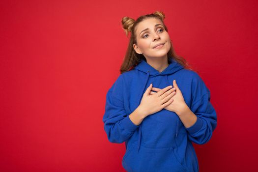 Portrait of young happy positive cute attractive blond woman with two horns with sincere emotions wearing stylish clothes isolated on red background with free space and holding hands on chest.