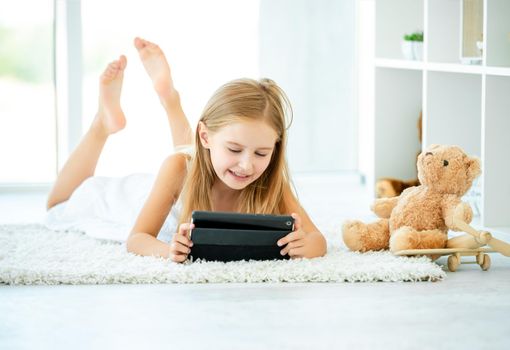 Kid girl lying on carpet in her room, watching video on digital tablet, near teddy bear