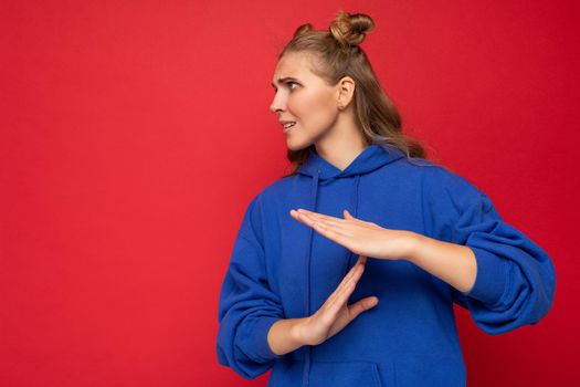 Photo of emotional young pretty blonde female person with two horns with sincere emotions wearing hipster bright blue pullover isolated over red background with copy space and showing timeout gesture.