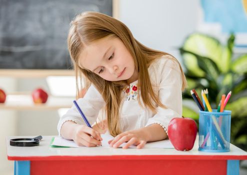 Little blond girl sitting at the white desk and writing classwork in the copybook in the school classroom
