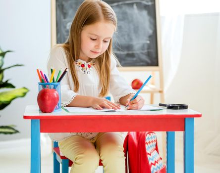 Little blond girl sitting at the white desk and writing classwork in the copybook in the school classroom