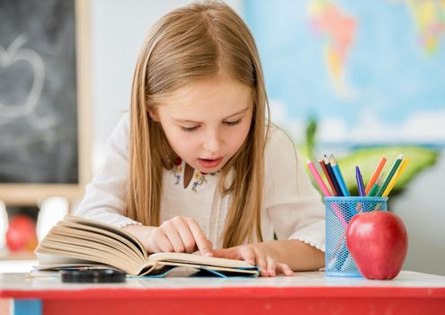 Little pretty blond girl sitting in the classroom at the white desk and attentively reading book