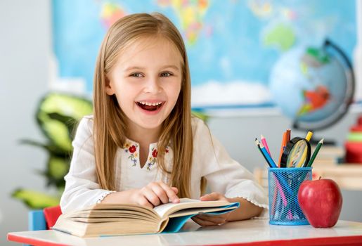 Little pretty blond girl sitting in the classroom at the white desk and reading book pointing by finger