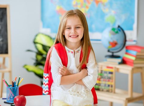 Little smiling blond girl standing near desk with her red bag in the spacious school class