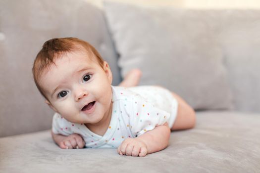 Cute little child in T-shirt lying on gray couch and looking at camera with opened mouth