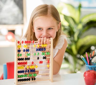 Little smiling blond girl sitting at the white desk and counting on the colourful abacus in the school classroom