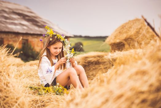 LIttle girl in white shirt sitting on a haystack and making a flower wreath