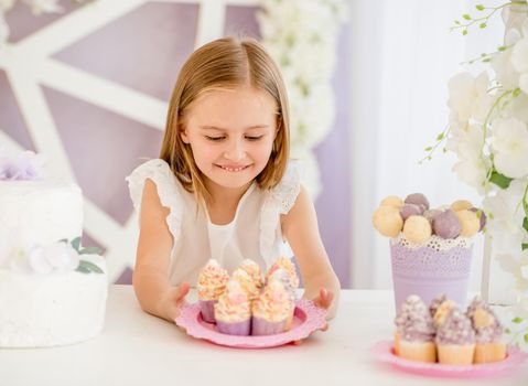Little pleasured blond girl holding a pink plate looking on sweet cakes on the background of pretty decorated candy bar