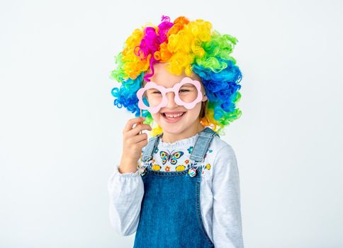 Cheerful schoolchild in rainbow wig holding stick with glasses for photo