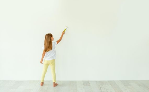 Little girl painting on white wall indoors. view from the back