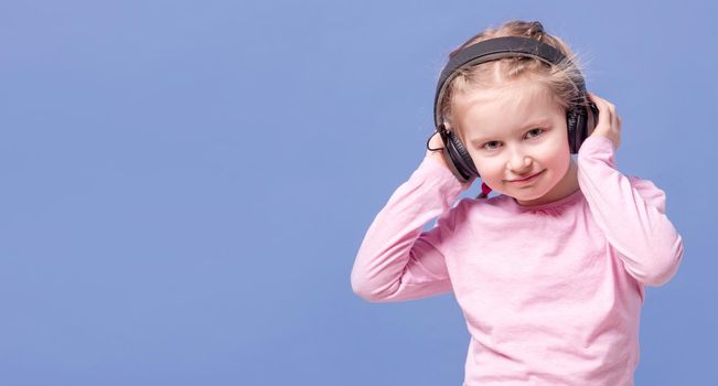 Cute little girl with headphones listening to music on light blue background. Copyspace