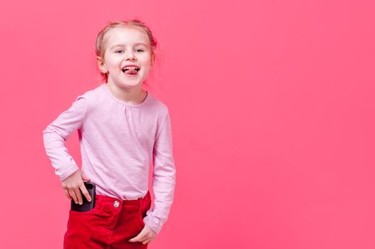 Cheerful little girl shows tongue while puts her cellphone in pocket, bright pink background