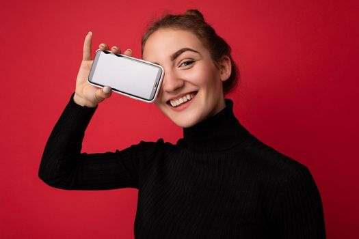 Photo of beautiful smiling young woman good looking wearing casual stylish outfit standing isolated on background with copy space holding smartphone showing phone in hand with empty screen display for mockup looking at camera.