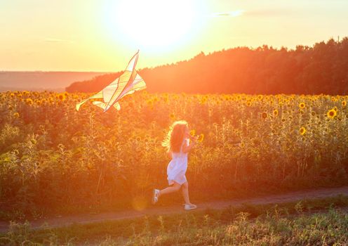 little cute girl in a white dress flying a striped kite in a meadow at sunset