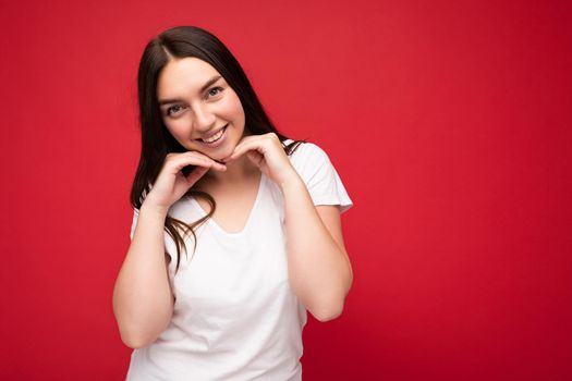 Photo of positive smiling young cute beautiful brunette female person with sincere emotions wearing casual white t-shirt for mockup isolated over red background with copy space.