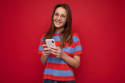 Photo shot of attractive positive good looking young woman wearing casual stylish outfit poising isolated on background with empty space holding in hand and using mobile phone messaging sms looking at camera.