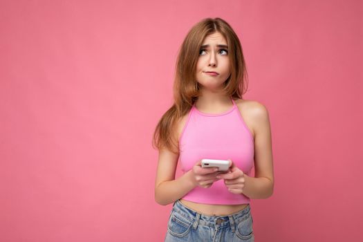 Beautiful thoughtful young blonde woman wearing pink top poising isolated on pink background with empty space holding in hand and using mobile phone surfing on the internet online looking to the side and having doubts. copy space