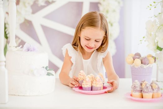 Little pleasured blond girl holding a pink plate looking on sweet cakes on the background of pretty decorated candy bar