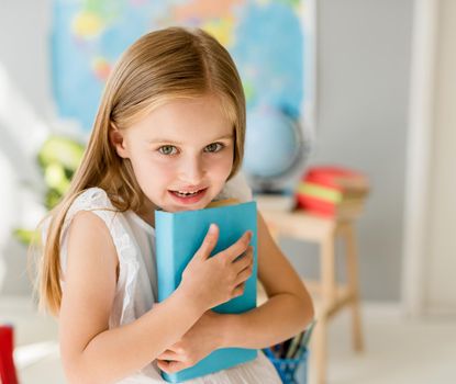 Little smiling blond girl sitting on the white desk and holding blue book in the sunshine school class