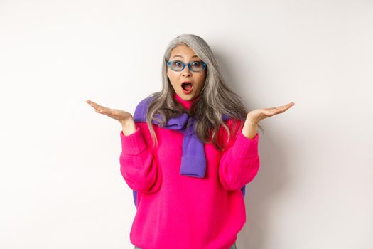 Fashionable asian senior woman in sunglasses gasping amazed, spread hands sideways and staring surprised at camera, white background.