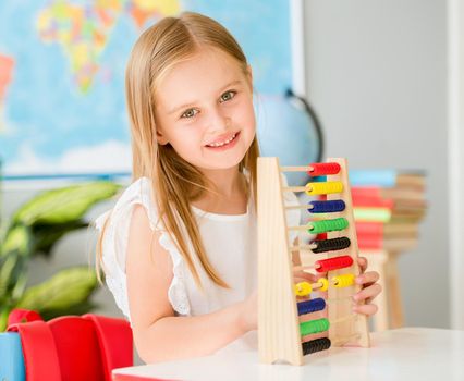 Little smiling blond girl sitting at the white desk and counting on the colourful abacus in the school classroom