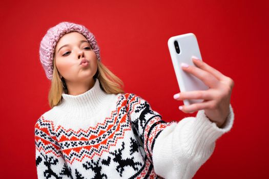 Photo of pretty positive young blonde woman wearing warm knitted hat and winter warm sweater standing isolated over red background using mobile phone taking selfie photo looking at device display and gicing kiss.