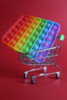 The multi-colored toy is popit simple dimple anti-stress in a basket of stroller shopping carts on a red background with a place for text copyspace. Shopping.