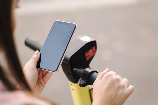 Young woman connect an electric scooter outdoors. Woman using phone for contactless payment. Ecological transportation concept.