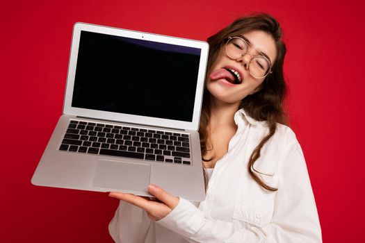 Crazy mad beautiful brunette curly young woman holding computer laptop wearing glasses white shirt showing tongue looking to the side isolated over red wall background. Empty space, cutout