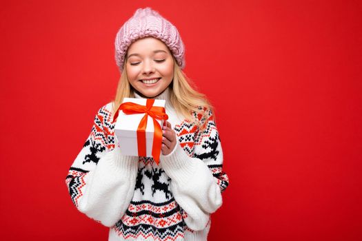 Photo of beautiful happy smiling young blonde woman isolated over red background wall wearing winter sweater and pink hat holding white gift box with red ribbon and looking at present. Copy space, mockup