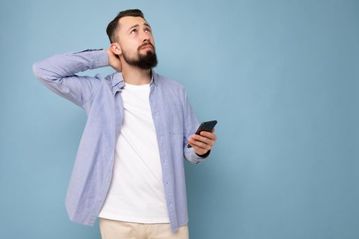 Thoughtful handsome young brunette unshaven man with beard wearing stylish white t-shirt and blue shirt isolated over blue background with empty space holding in hand and using phone messaging sms looking up. Copy space