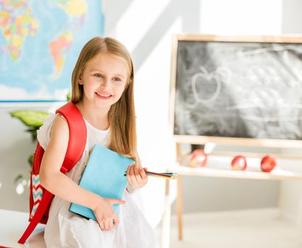 Little smiling blond girl standing near blackbord with red backpack and holding a notebook with pencils in the sunshine school classroom