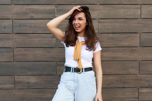 Portrait of successful smiling joyful happy young brunet woman wearing casual white t-shirt and jeans with yellow sweater poising near brown wall in the street and having fun.