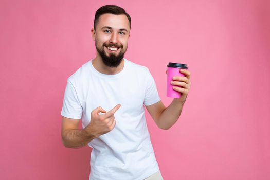 photo of handsome happy young brunette unshaven man with beard wearing white t-shirt isolated over pink backgroung wall holding paper coffee cup drinking and looking at camera and pointing finger at beverage.