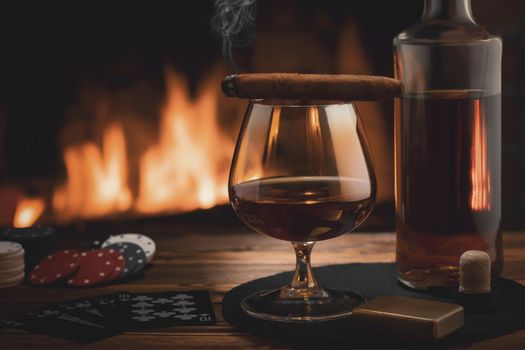 Glass of cognac, cigar, bottle, cards game and chips on the table near the burning fireplace. Rest, pleasure, good luck and dolce vita concept.