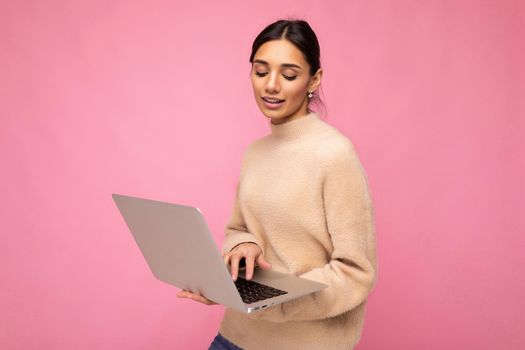 Close-up portrait of Beautiful young brunette woman wearing beige sweater holding netbook computer typing text on keyboard looking down isolated over pink wall background.