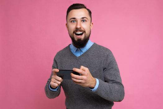 Shot of happy handsome young brunette unshaven man with beard wearing everyday grey sweater and blue shirt isolated on background wall holding smartphone playing games via mobile phone looking at camera and having fun.