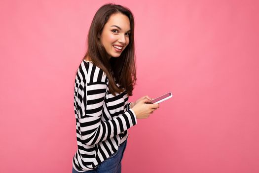 Beautiful young brunette woman using mobile phone communicating via texting message wearing sweater isolated on wall background looking at camera.