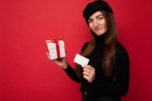 Attractive positive young brunette woman wearing black sweater and hat isolated on red background holding credit card and present box looking at camera. copy space