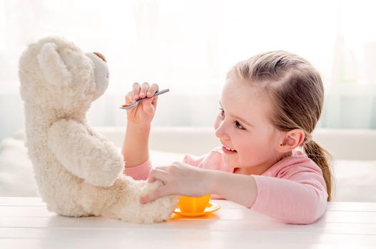 Cute little girl feeding her teddy bear with spoon. Beautiful child girl sitting at a table and feed teddy bear. Happy little kid playing with toy teddy bear
