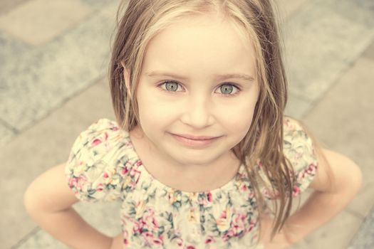 Sweetie blonde girl in dress with flower pattern looks at camera and smiling shyly.