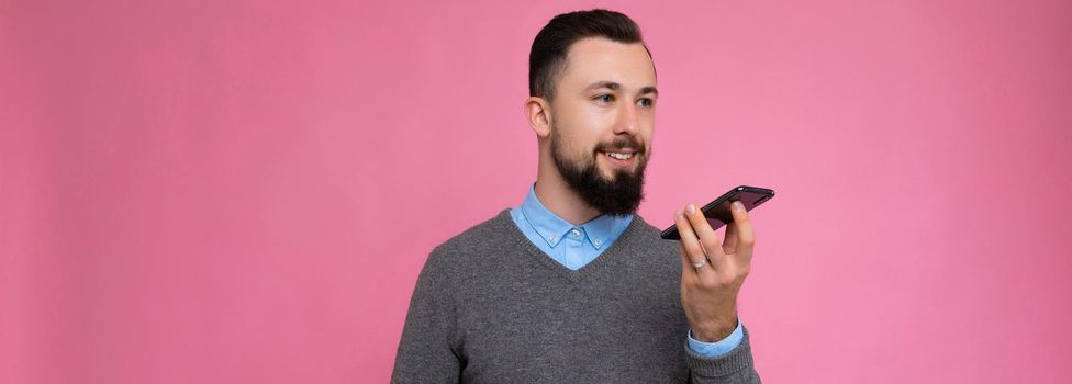 Panoramic photo of handsome positive good looking young man wearing casual stylish outfit poising isolated on background with empty space holding in hand and using mobile phone recording voice message looking at smartphone display screen.