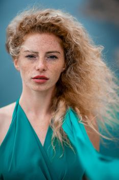 Outdoor portrait of a young beautiful natural redhead girl with freckles, long curly hair, in an emerald dress, posing against the background of the sea