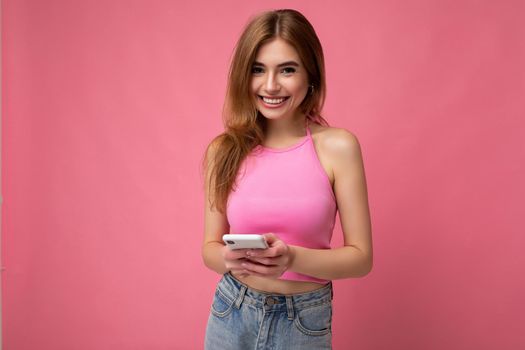 Photo of good looking smiling young blonde woman wearing pink top poising isolated on pink background with empty space holding in hand and using mobile phone messaging sms looking at camera. copy space