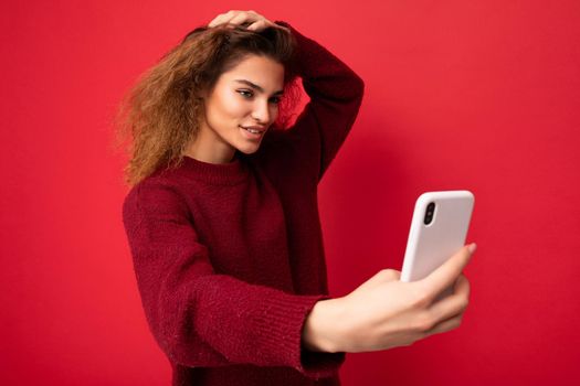 Shot of sexy smiling Beautiful young woman with curly hair wearing dark red sweater isolated on red background wall holding and using smart phone looking at telephone screen and taking selfie.