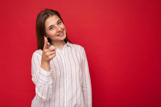 Young smiling beautiful brunette woman with sincere emotions isolated on background wall with copy space wearing casual white shirt and pointing finger at camera. Positive concept.