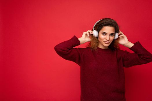 Beautiful happy smiling young brunette curly woman wearing dark red sweater isolated over red background wall wearing white bluetooth headphones listening to music and having fun looking at camera. copy space
