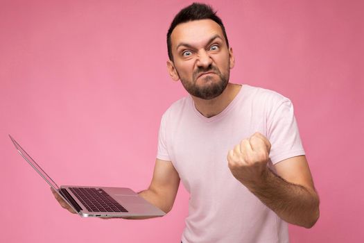 Handsome angry brunet man holding laptop computer showing fist looking at camera in t-shirt on isolated pink background.