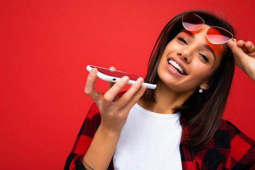 Photo shot of smiling attractive positive good looking young woman wearing casual stylish outfit poising isolated on background with empty space holding in hand and using mobile phone recording voice message looking at camera.