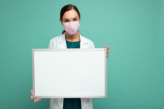 Young attractive female doctor in protective face mask and white medical coat holding an empty magnetic board isolated on blue background.